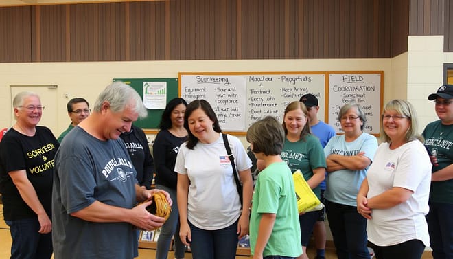 A group of smiling parents gathered in a well-lit school gymnasium, dressed casually and ready to volunteer. In the foreground, a parent helping a child with a baseball glove, while in the middle, others are setting up equipment and organizing supplies. The background features a bulletin board with handwritten signs advertising various support roles - scorekeeping, equipment management, field maintenance, and team coordination. The overall atmosphere exudes a warm, community-driven spirit, as parents enthusiastically contribute their time and effort to enrich the young baseball players' experience.
